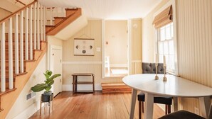 A spacious room with a wooden staircase, a dining table with chairs, and a large window providing natural light.