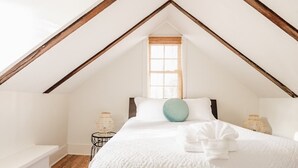 A bedroom with a sloped ceiling, a bed with white bedding, a window with blinds, and two wicker lamps.