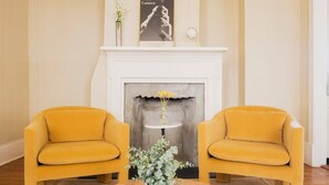 A room with two yellow armchairs facing a fireplace, a small table with a vase of flowers, and a poster above the fireplace. - Historic Charleston Charm (Charleston)