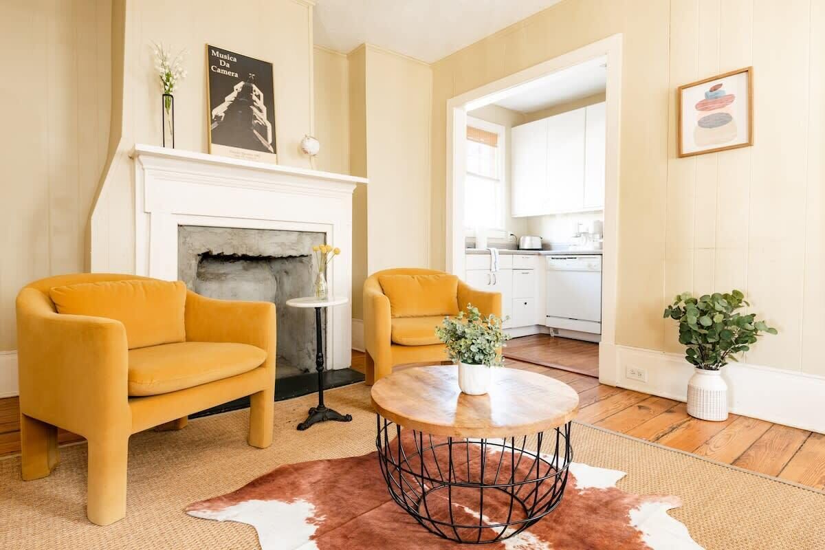 A living room with two yellow armchairs, a round wooden coffee table, a fireplace, and a kitchen in the background.