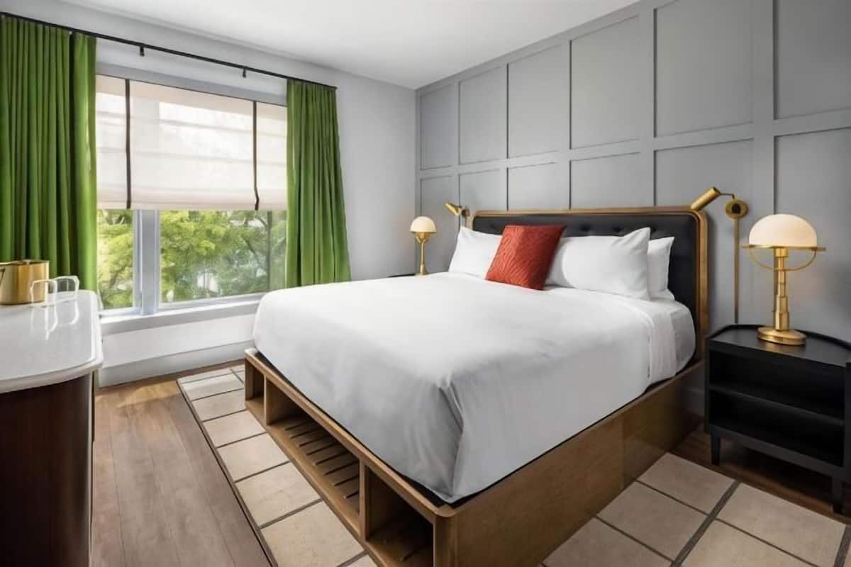 A hotel room with a bed, bedside tables, a desk, a window with curtains, and a view of greenery outside.