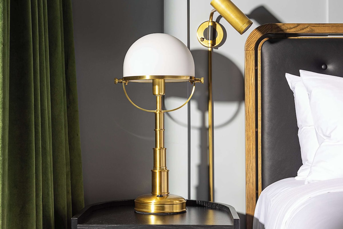 A neatly made bed with white linens, a gold-toned floor lamp with a white shade, and a dark wooden nightstand with a black marble top.
