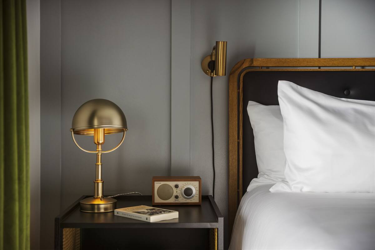A neatly made bed with white linens, a wooden headboard, a bedside table with a vintage radio, and a classic lamp.