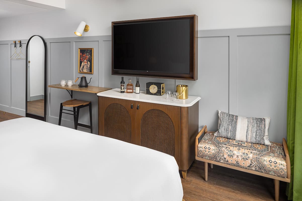 A hotel room with a bed, a desk with a flat-screen TV, a wooden cabinet with bottles, a chair, and a patterned sofa.