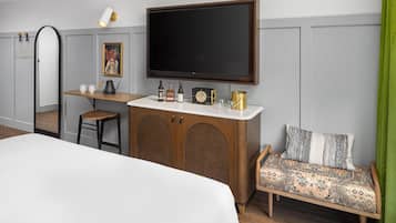 A hotel room with a bed, a desk with a flat-screen TV, a wooden cabinet with bottles, a chair, and a patterned sofa.
