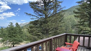 Eine Holzterrasse mit roten Stühlen und einem Tisch, mit Blick auf eine gebirgige Landschaft mit Bäumen und blauem Himmel.