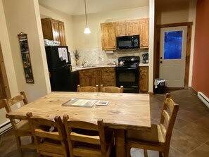 A compact kitchen with wooden cabinets, a black refrigerator, a microwave, and an oven. There is a dining table with chairs and a hanging light fixture. - Bears Den 2- Mountain Duplex, Views, Wildlife & Hot Tub (Estes Park)