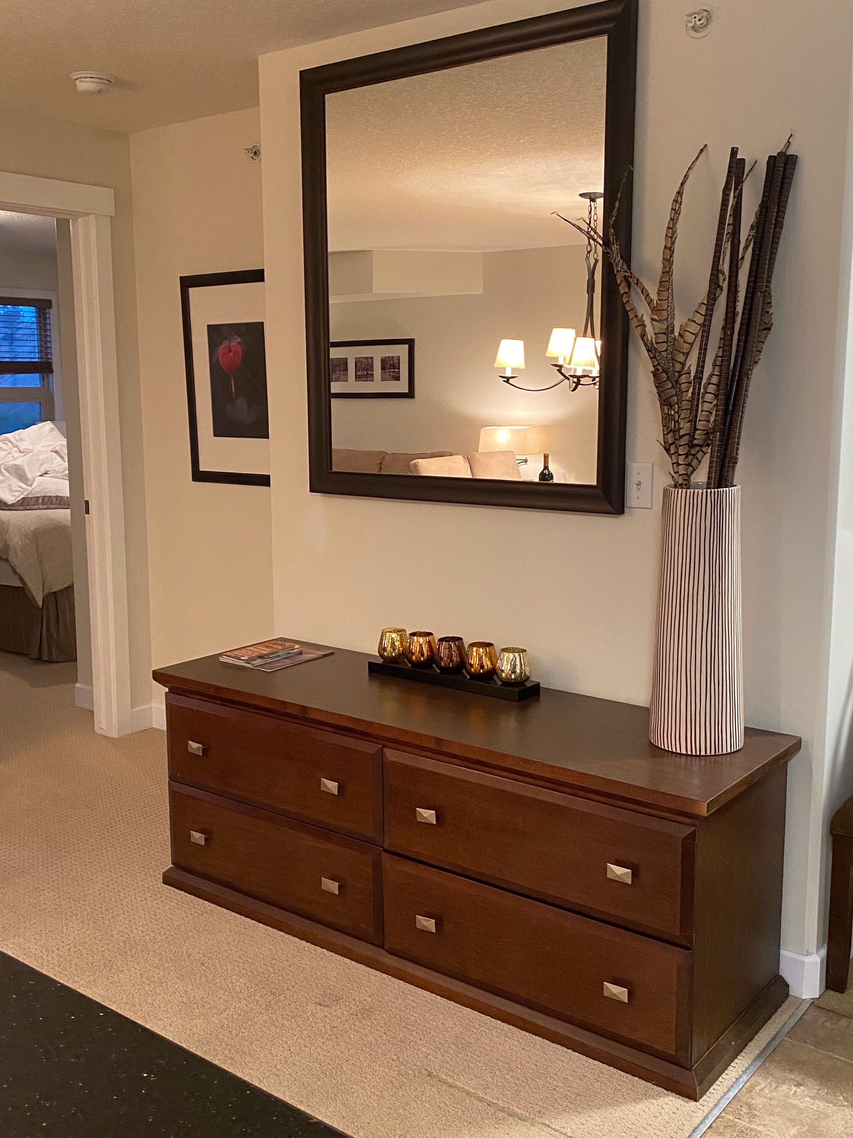 Une commode en bois avec un miroir au-dessus, un vase avec des branches et un tableau encadré accroché au mur.