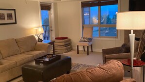A living room with a sofa, ottoman, and chairs, a coffee table, a lamp, and a window with a view. - Top floor condo on Lake Windermere with spectacular lake and Mountain View’s (Invermere)