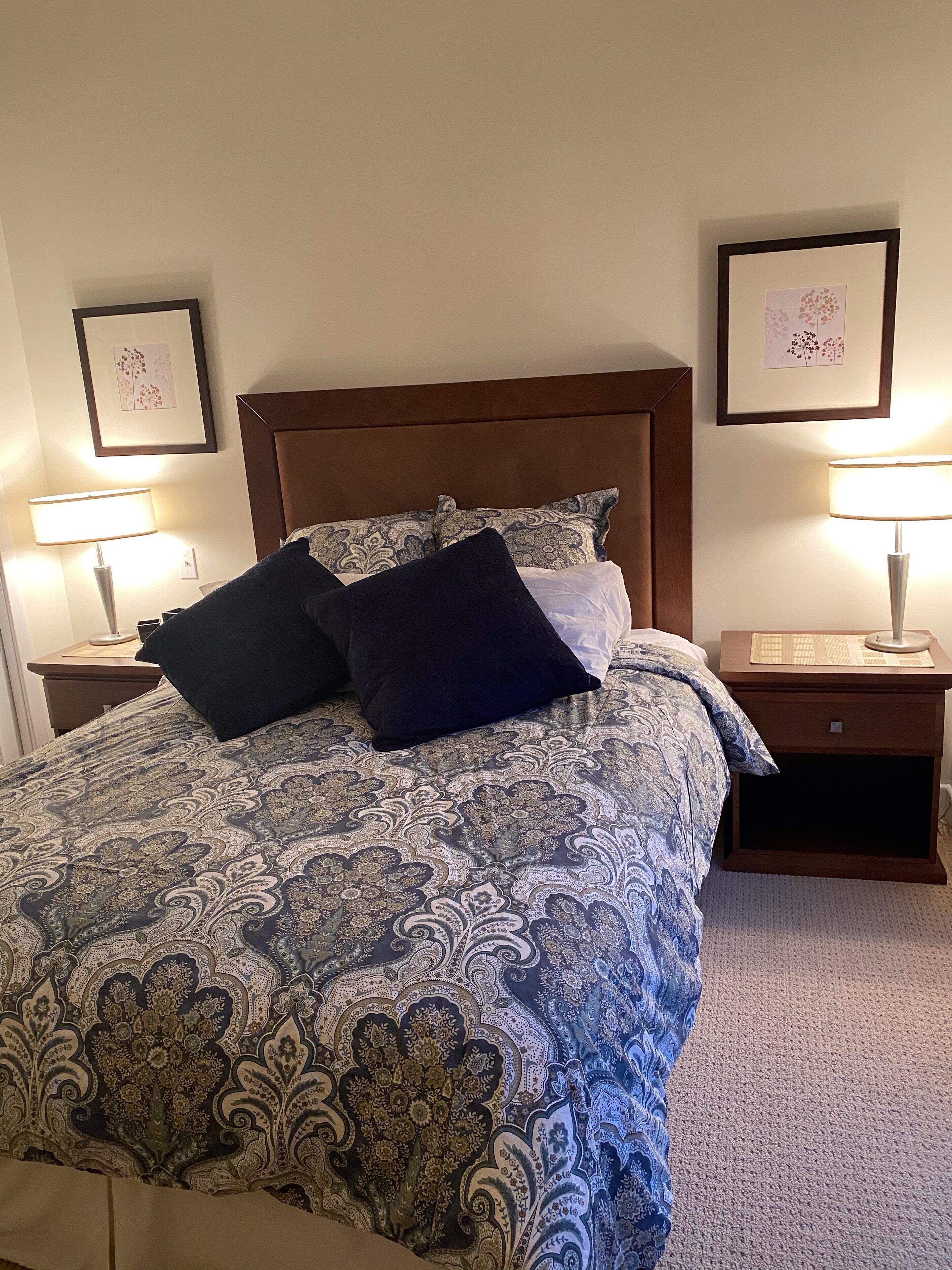A neatly made bed with a patterned bedspread, two pillows, and a wooden headboard. There are two bedside tables with lamps, and framed pictures on the walls.