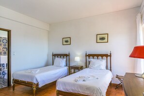 Ein Hotelzimmer mit zwei hölzernen Betten, weißer Bettwäsche, einem kleinen Nachttisch mit Lampe und zwei gerahmten Bildern an der Wand.