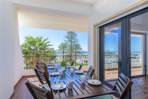 A balcony with a dining table set for four, overlooking a coastal view with palm trees and a clear sky.