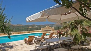 Zona de piscina exterior amb gandules i una taula sota un para-sol gran, envoltada de vegetació i amb vistes a les colines llunyanes.