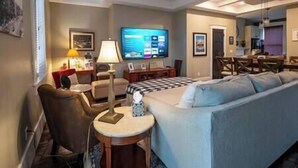 A hotel room with a sofa, armchair, small table, lamp, TV, and a kitchenette area.