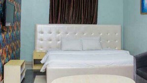 A bedroom with a white bed, a small bedside table, a chair, a round table, and a colorful wall. - Room-ambience Hotel Magodo - Guest Room (Lagos)