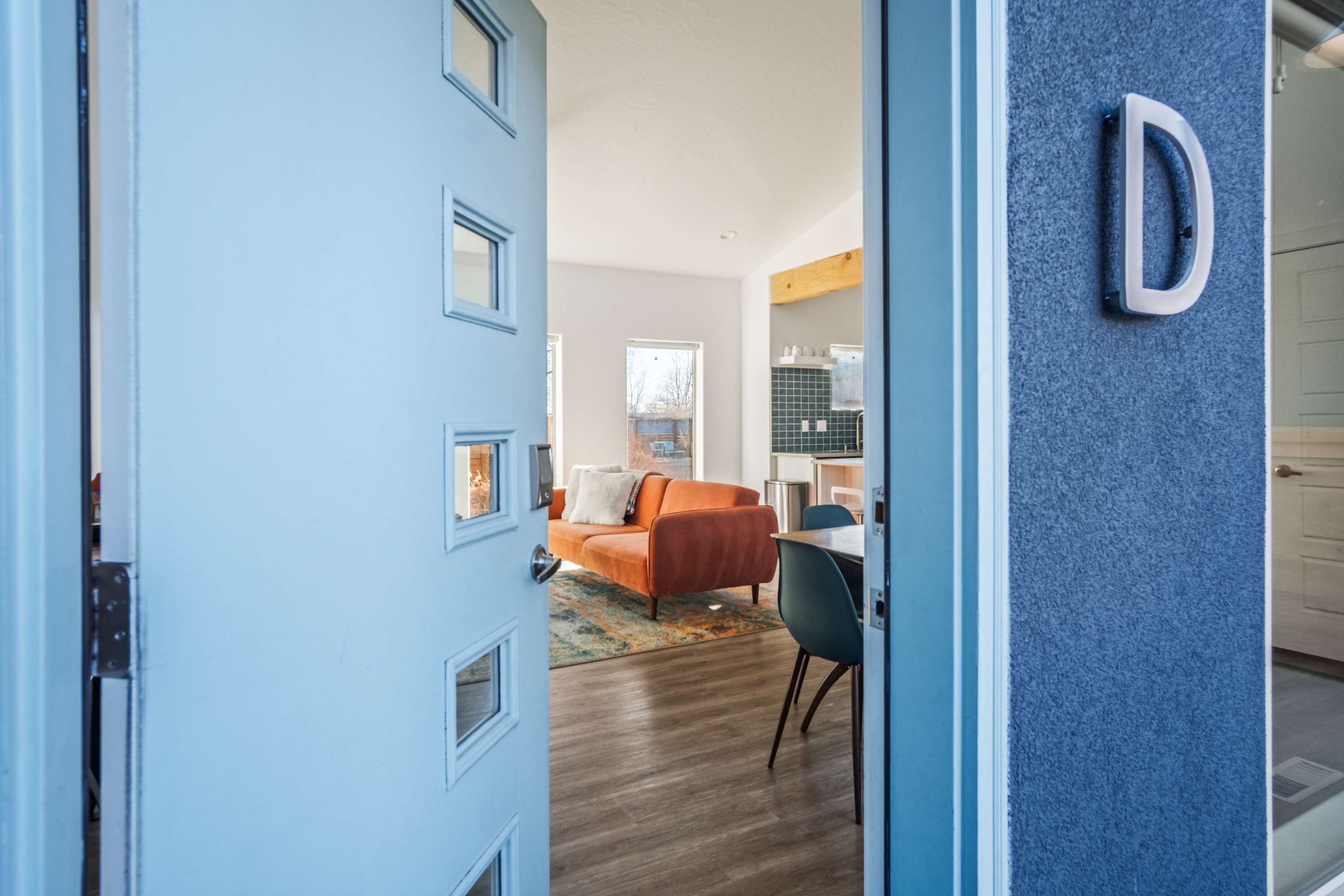 A modern interior with a blue door, a living area with a sofa and a dining table, and a kitchen area with a tiled backsplash.