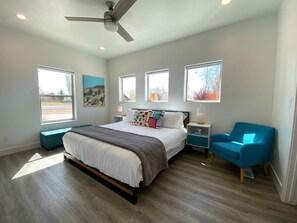 A bedroom with a bed, a ceiling fan, a nightstand, a blue chair, and a window with a view of the outside.
