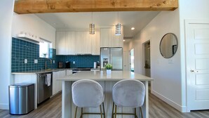 A modern kitchen with a central island, two bar stools, a stainless steel trash bin, and a wooden ceiling beam.