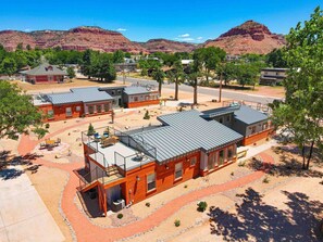 A modern building with a flat roof and a balcony, surrounded by a landscaped area with trees and a clear view of red mountains in the background.