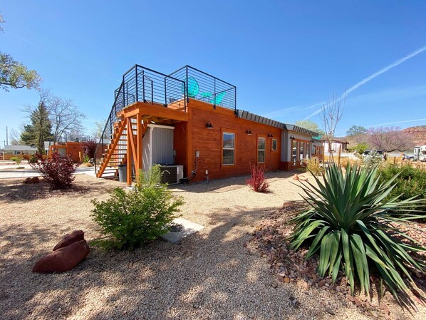 A modern wooden building with a metal staircase, a covered outdoor area, and a gravel driveway.