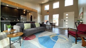 Una sala moderna con un sofá gris, un sillón rojo y una mesa de centro. Se observa una zona de cocina con armarios oscuros y electrodomésticos de acero inoxidable. La habitación cuenta con grandes ventanales que permiten la entrada de luz natural.