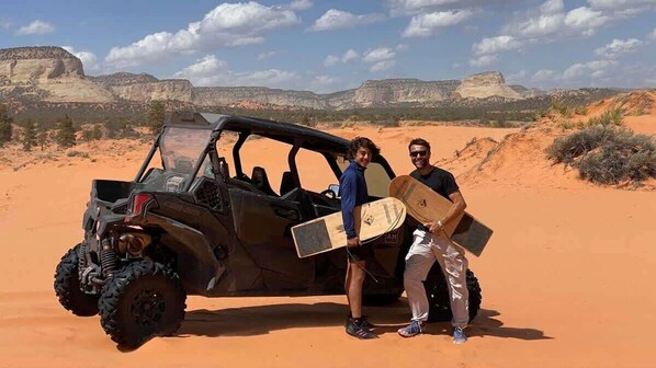 A desert landscape with a vehicle and two people holding surfboards.