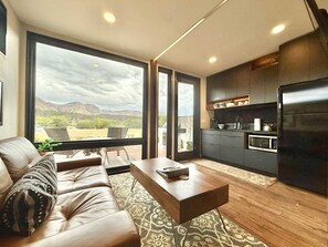 A modern living room with a leather sofa, a coffee table, and a view of a mountainous landscape through large windows. - East Zion Designer Container Studio- The Fields (Orderville)