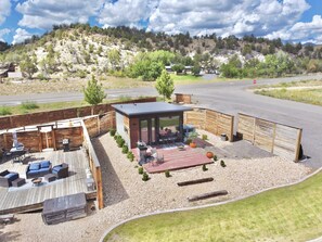 A modern outdoor patio with seating and a barbecue area, surrounded by a wooden fence and a view of a road and hills. - East Zion Designer Container Studio- The Fields (Orderville)