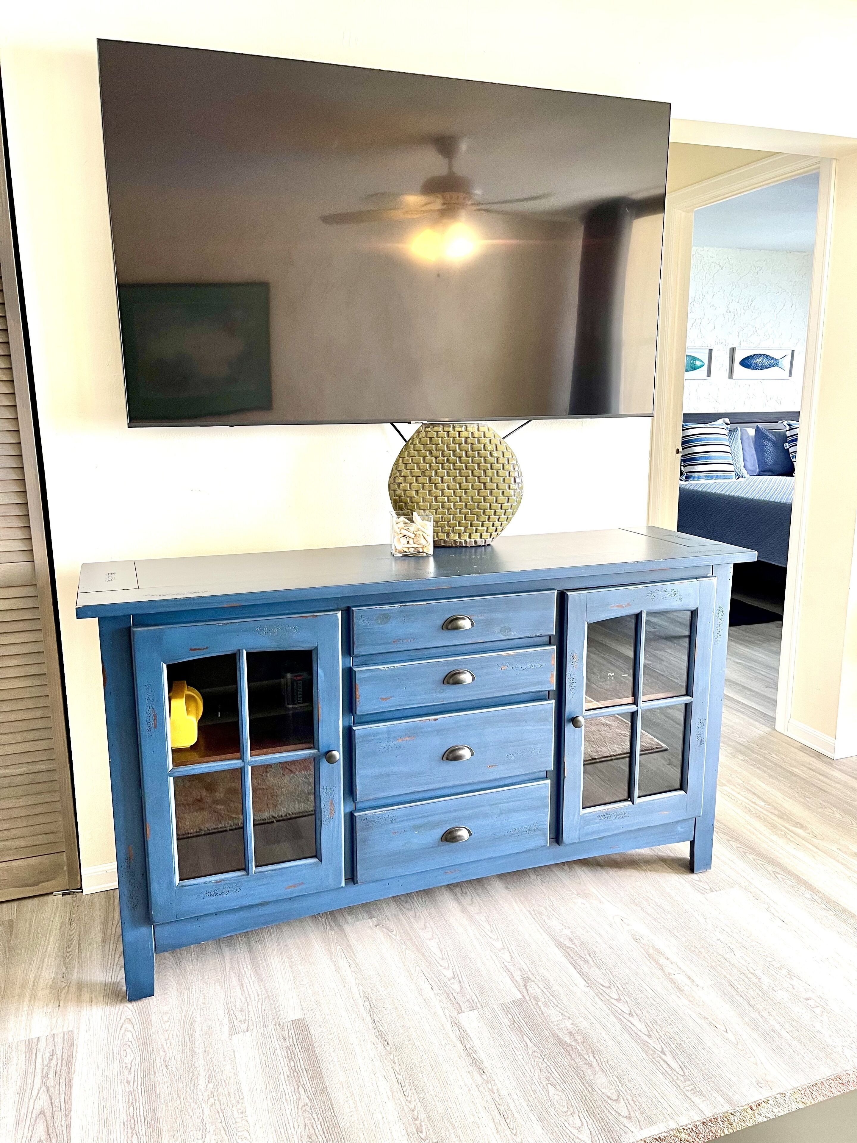 A blue wooden cabinet with drawers and a glass door, placed in a room with a large flat-screen TV mounted on the wall.