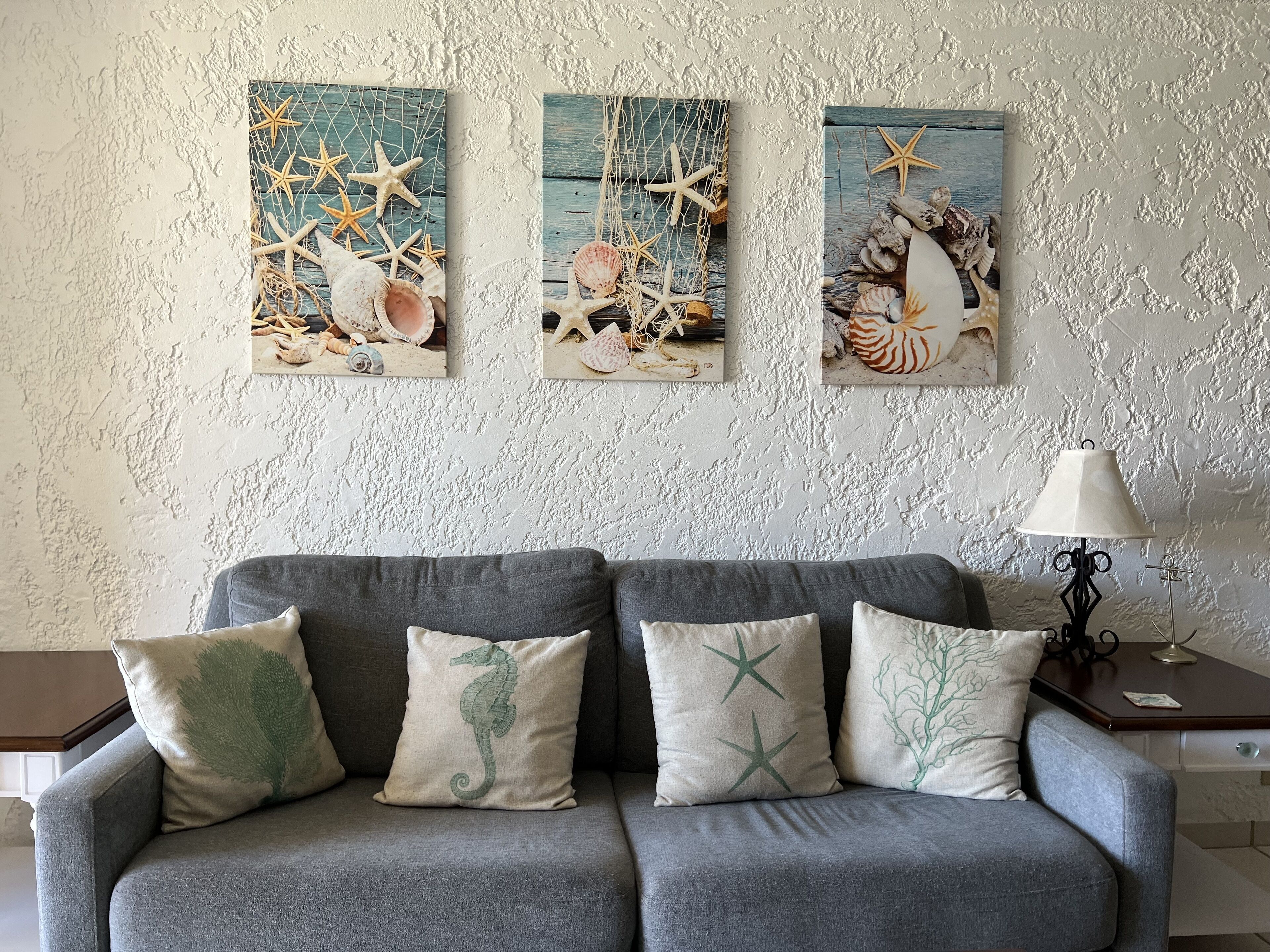 Un canapé gris avec trois coussins, une lampe et trois tableaux accrochés au mur, représentant des motifs marins.