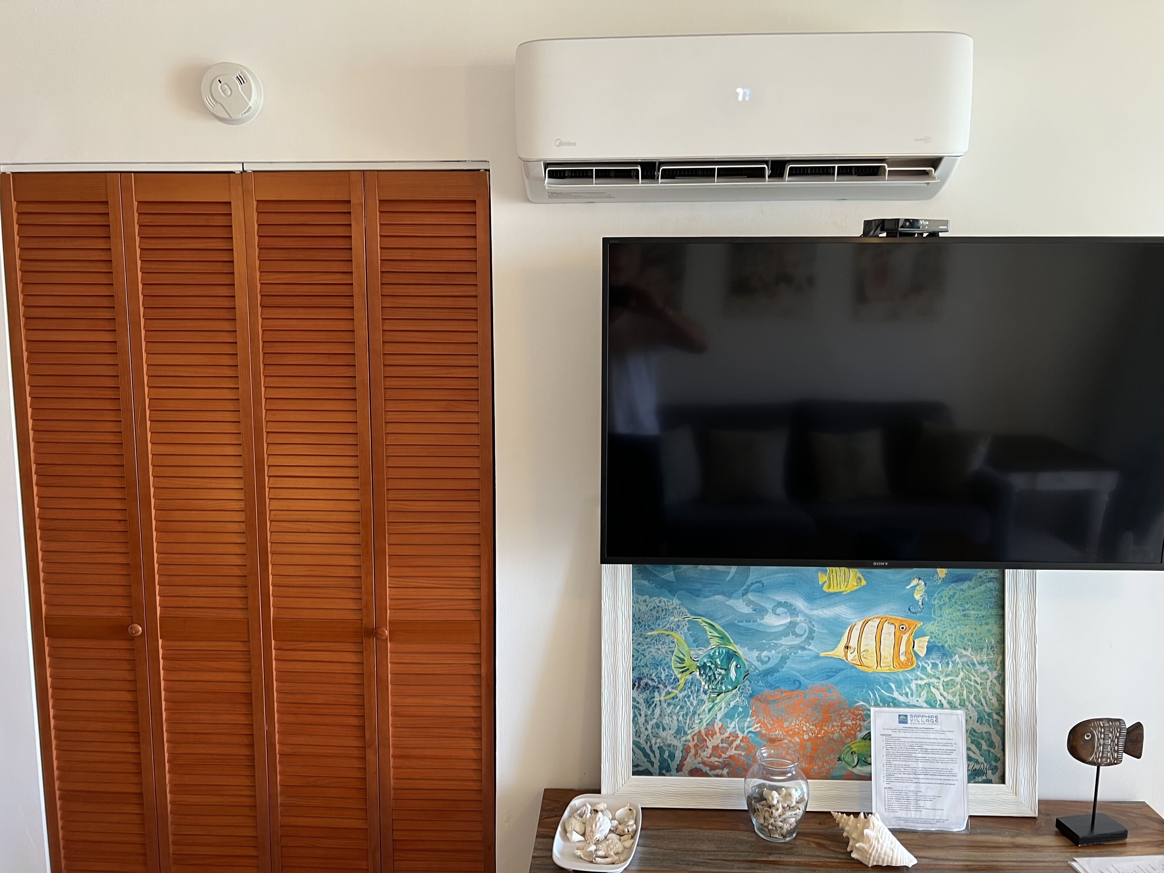 A room with a wooden closet, a mounted air conditioner, a flat-screen TV, and an aquarium.