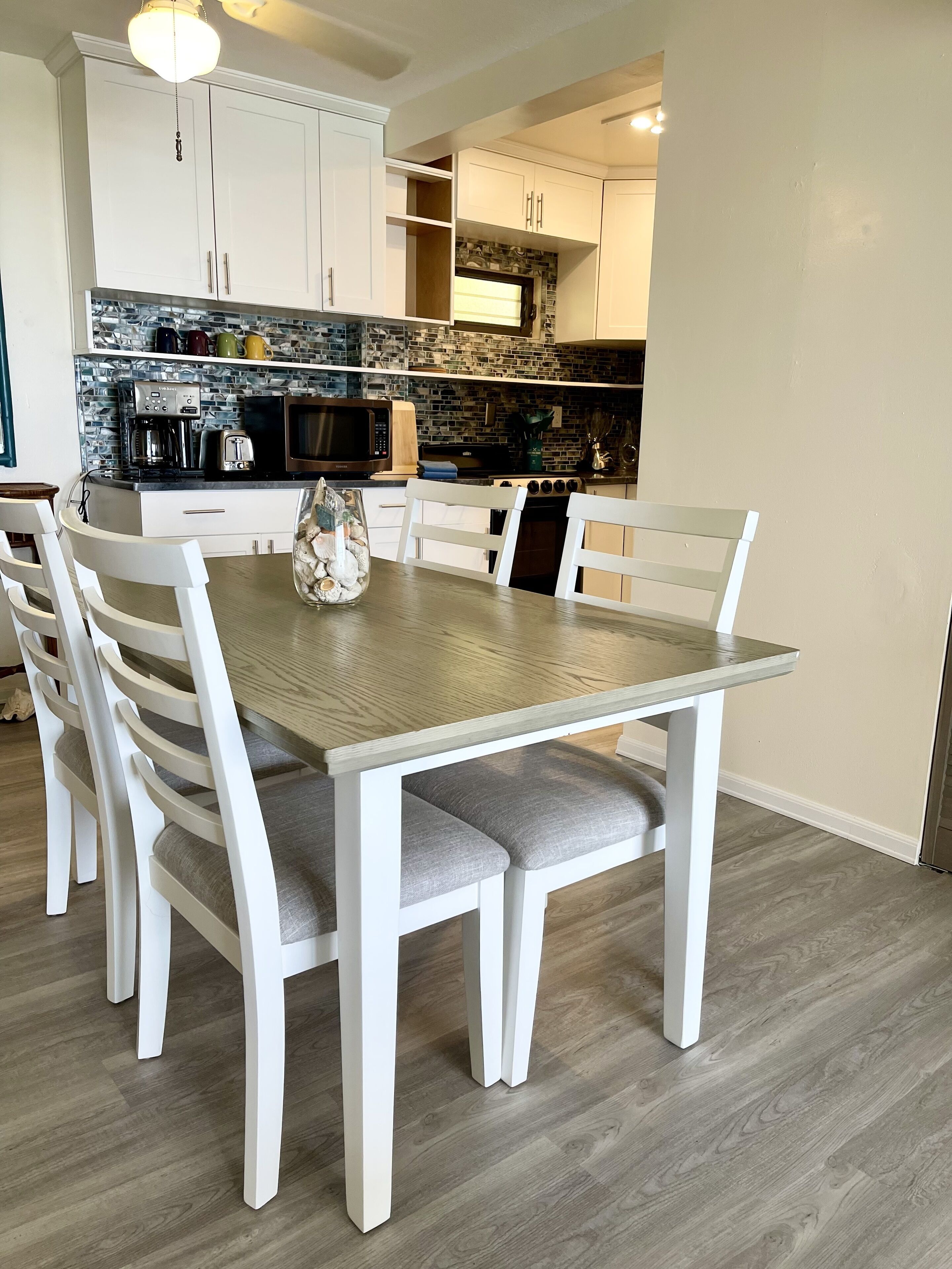 Un comedor con una mesa rectangular y seis sillas blancas, una cocina al fondo con armarios blancos y una encimera negra, y piso de madera.