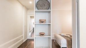 A hallway with a white wall, wooden floor, and a bed with a white mattress and brown blanket. There are three shelves with baskets and a remote control. - GuestReady - Bourse Maison (Porto)
