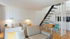 A modern living room with a sofa, patterned cushions, and pendant lights. A staircase leads to a kitchen area with a dining table and chairs.