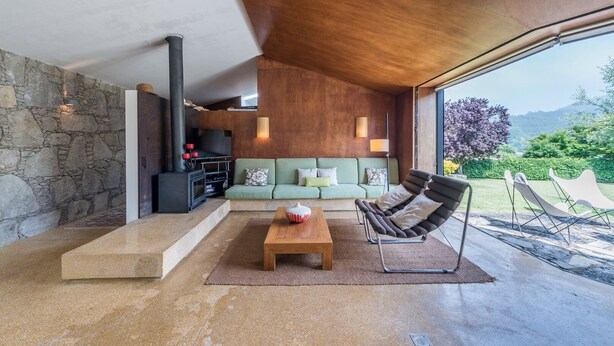 A modern living room with a stone wall, a wooden ceiling, a green sofa, a wooden coffee table, and a fireplace. - GuestReady - Chapel`s Garden (Caminha)