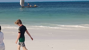 Escena de playa con un velero en el agua y gente caminando sobre la arena.
