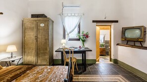 A compact room with a wooden cabinet, a small table with a lamp, a chair, a bed with a patterned bedspread, a television on a wooden stand, and a window with a curtain. - Authentic Sicilian Charm With Pool, Sea View, Parking & Wifi (Avola)