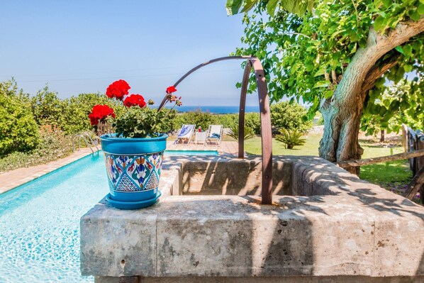 A poolside area with a blue ceramic pot with red flowers, a wooden pergola, and a view of the sea. - Authentic Sicilian Charm With Pool, Sea View, Parking & Wifi (Avola)