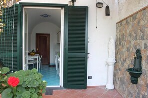 An open door leading to a dining area with a table and chairs, a stone wall with a fountain, and a white statue. - Eden Holiday Apartment (Minori)
