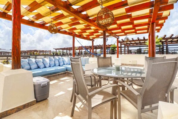 An outdoor patio with a round glass table and chairs, a blue sofa with striped cushions, and a wooden pergola overhead.