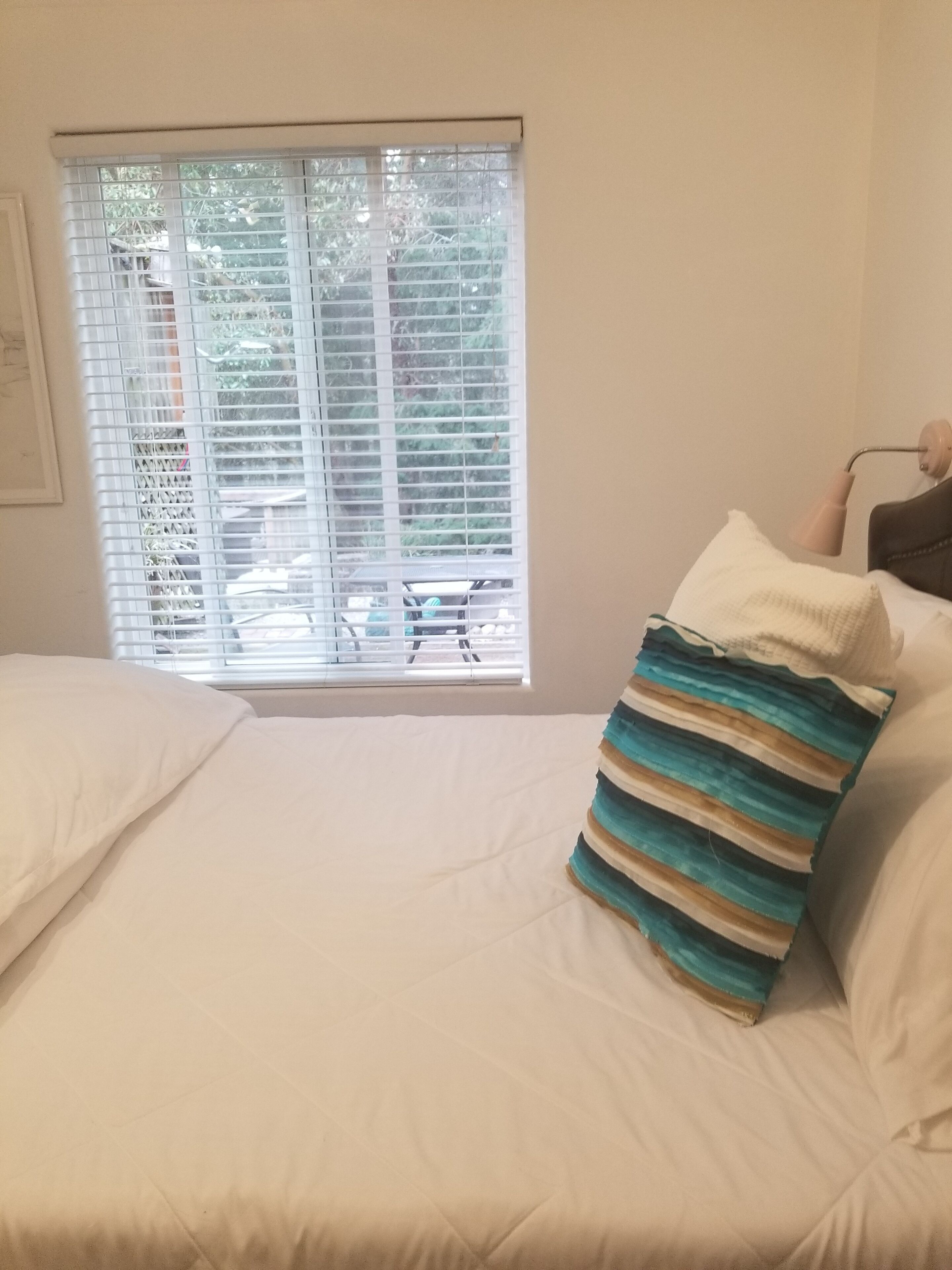Un dormitorio con una cama, almohadas blancas, una almohada a rayas, una ventana con persianas y una lámpara en una mesita de noche.