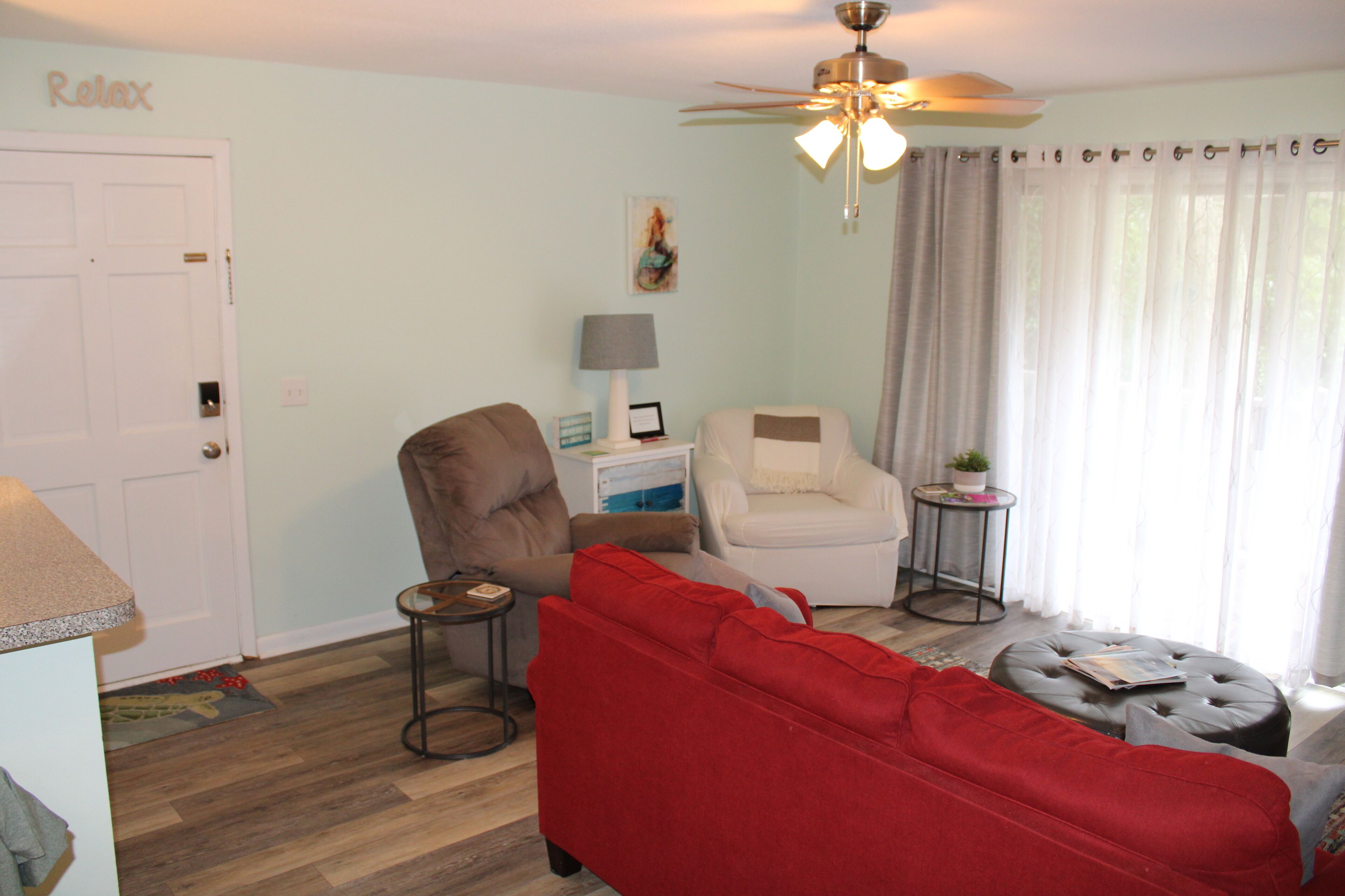 A living room with a red sofa, a white armchair, a brown armchair, a ceiling fan, and a small round table with a lamp.