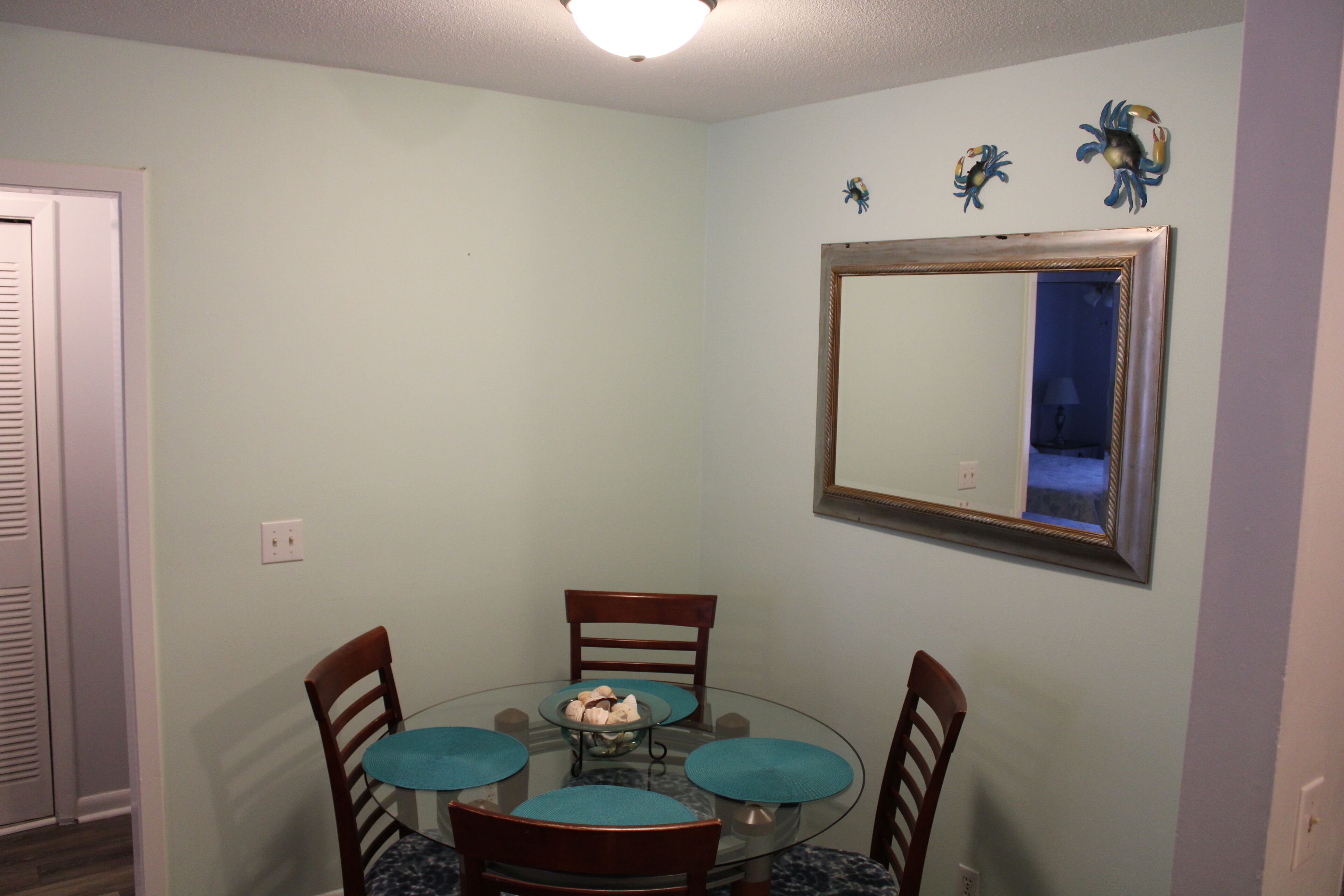 Un comedor con una mesa redonda de vidrio, cuatro sillas de madera con cojines azules, y un espejo colgado en la pared sobre la mesa.