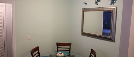 Un comedor con una mesa redonda de vidrio, cuatro sillas de madera con cojines azules, y un espejo colgado en la pared sobre la mesa.