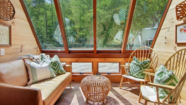 A cozy cabin interior with a wooden floor, a sofa, two chairs, a woven ottoman, and a large window offering a view of a forest. - Hideaway at Harvest Moon (Sylva)