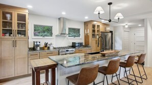 A modern kitchen with wooden cabinets, a stainless steel refrigerator, a large island with bar stools, and pendant lighting. - Hideaway at Harvest Moon (Sylva)