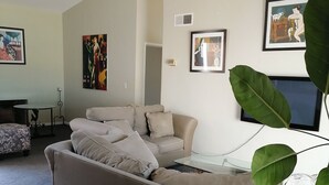 A modern living room with a large beige sectional sofa, a glass coffee table, and a flat-screen TV. There are several framed artworks on the walls and a large green plant in the foreground. - Ocean view beautiful apartament with pool and jacuzzi, furnished. (Tijuana y Rosarito)
