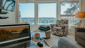 Quarto aconchegante com laptop, caneca e cadeira, próximo a uma janela grande com vista para um porto.