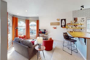Un salon moderne avec un canapé, un pouf rouge et un téléviseur. Il y a également un coin cuisine avec un bar et des tabourets.