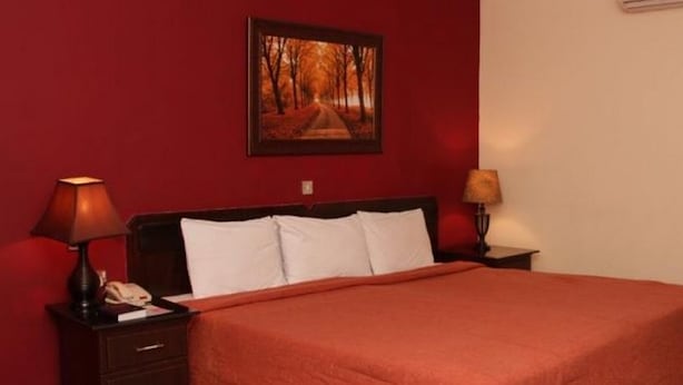 A hotel room with a large bed, two bedside tables with lamps, a nightstand, a framed picture on the wall, and a red carpet. - Room-agura Hotel - Guest Room (Abuja)